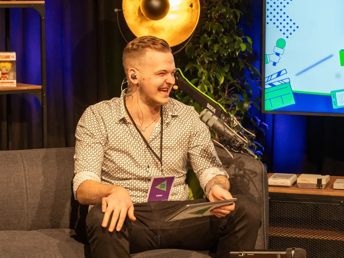 Das Bild zeigt einen Mann, der als Moderator namens Huebi beschrieben wird. Er sitzt auf einem Sofa, trägt ein Mikrofon und lächelt. Neben ihm ist ein Mikrofon auf einem Stativ und im Hintergrund ein großer Leuchtscheinwerfer.