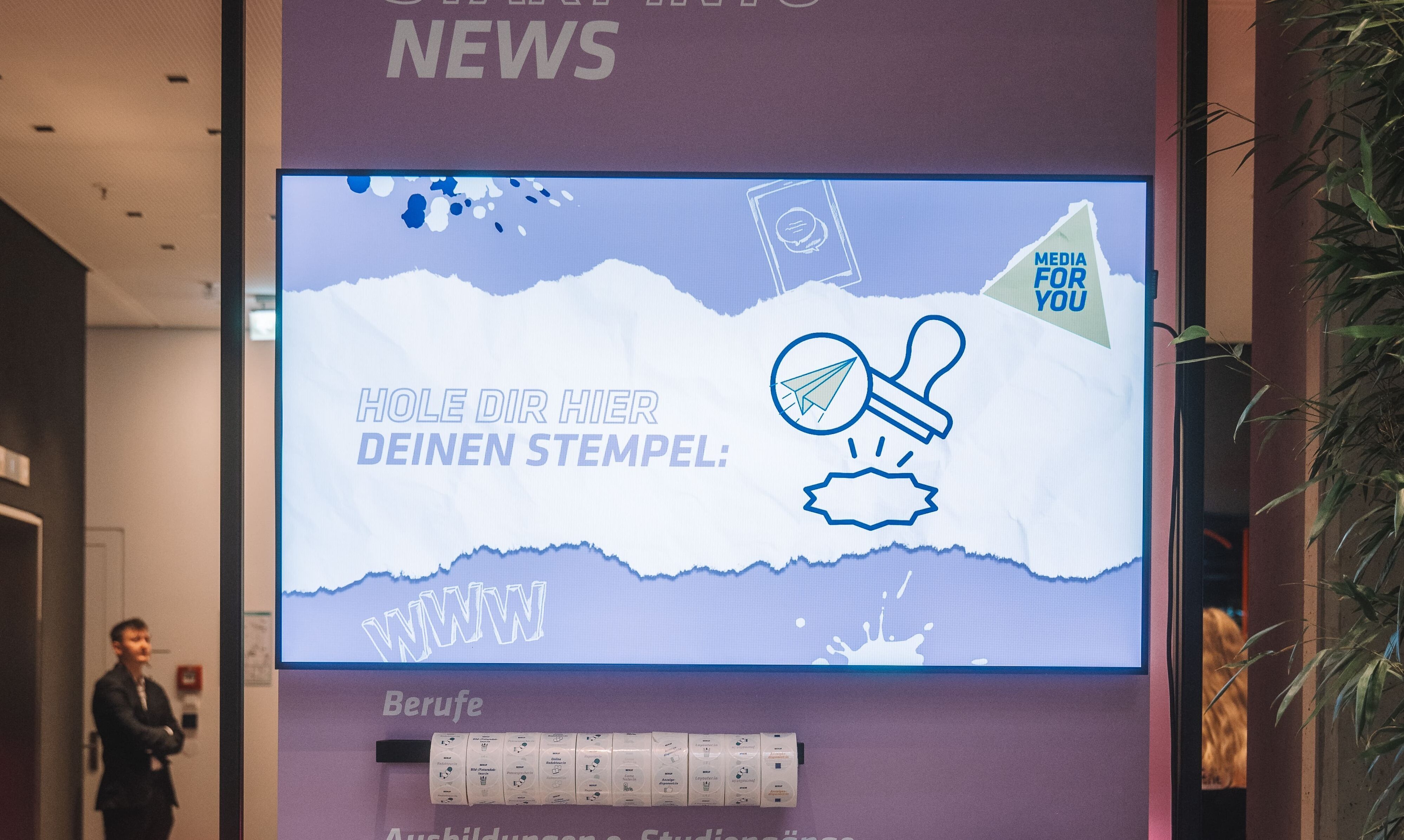 Großer Bildschirm mit Text „Hole dir hier deinen Stempel!“ und einem Stempel-Symbol, im Hintergrund stehen Personen.