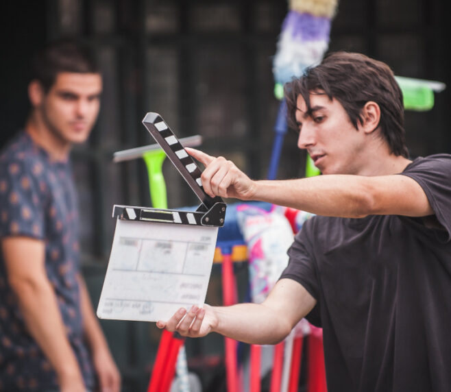 Ein junger Mann hält eine Filmklappe am Set und markiert den Beginn einer Szene, im Hintergrund steht eine weitere Person unscharf im Bild.