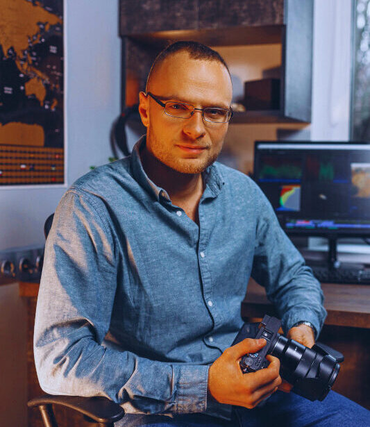 Ein Mann mit Brille sitzt in einem modernen Büro an einem Schreibtisch und hält eine Kamera in der Hand, im Hintergrund läuft ein Bildschirm mit Schnittsoftware.