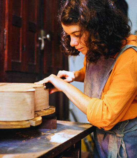 Eine Frau arbeitet in einer Werkstatt an einem hölzernen Musikinstrument, wie einem Cello oder einer Gitarre. Sie trägt ein orangefarbenes Hemd und eine Schürze und bearbeitet konzentriert die Oberfläche des Instruments. Im Hintergrund hängen Werkzeuge an der Wand.