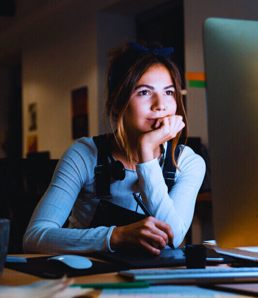 Junge Frau sitzt abends konzentriert vor einem Computer, stützt den Kopf in die Hand und arbeitet mit einem Grafiktablett.