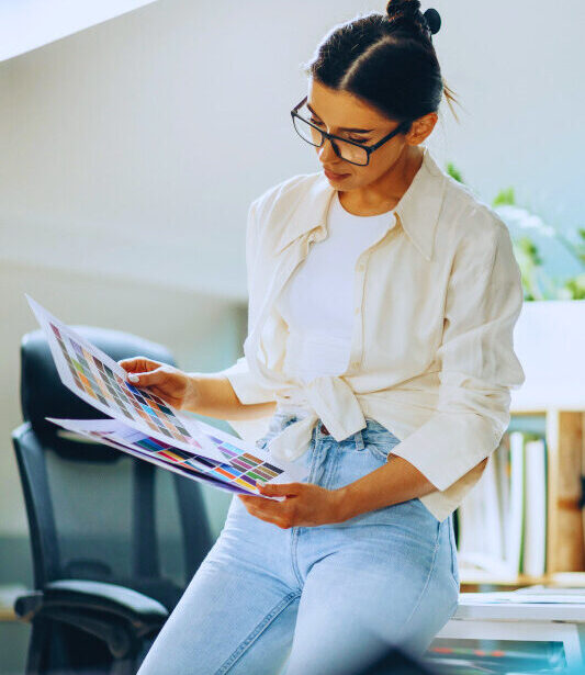Eine Frau mit Brille steht in einem modernen Büro und betrachtet aufmerksam Farbmuster auf zwei großen Blättern. Sie trägt ein weißes Hemd, ein T-Shirt und eine helle Jeans. Im Hintergrund sind ein Schreibtisch mit Computer, Regale und Pflanzen zu sehen.