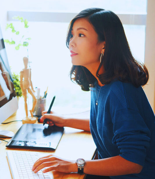 Eine Frau sitzt an einem Schreibtisch vor einem großen Computerbildschirm und arbeitet mit einem Grafiktablet. Auf dem Monitor sind bunte Bildcollagen zu sehen. Neben ihr stehen eine Lampe, Pflanzen und Zeichenutensilien. Tageslicht fällt durch ein Fenster in den Raum.