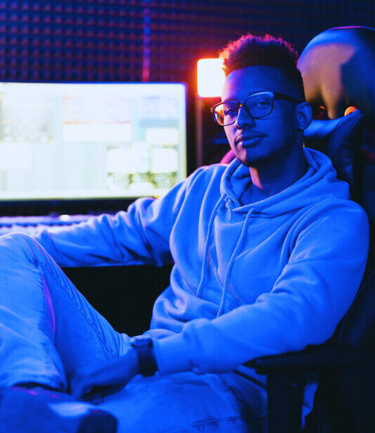 Ein junger Mann sitzt entspannt in einem Musik- oder Tonstudio auf einem Drehstuhl. Er trägt eine Brille und einen Kapuzenpullover, im Hintergrund ist ein großer Monitor mit Audioprogramm sowie Studiolautsprecher, Keyboard und Kopfhörer zu sehen. Das Licht im Raum ist in Blau- und Orangetönen gehalten und erzeugt eine kreative Arbeitsatmosphäre.