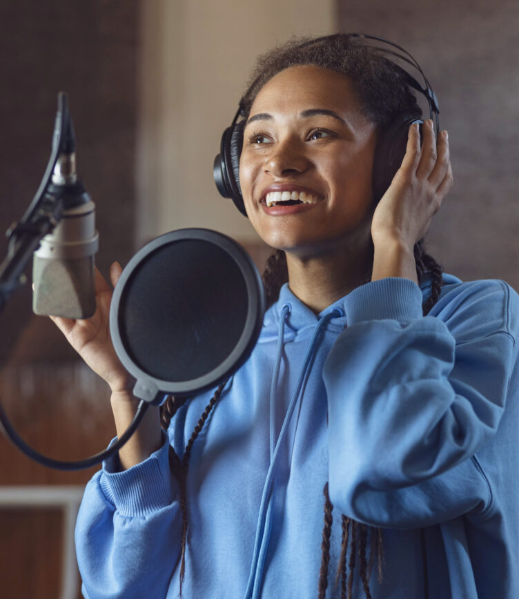 Eine junge Frau im Tonstudio trägt Kopfhörer und spricht fröhlich ins Mikrofon.