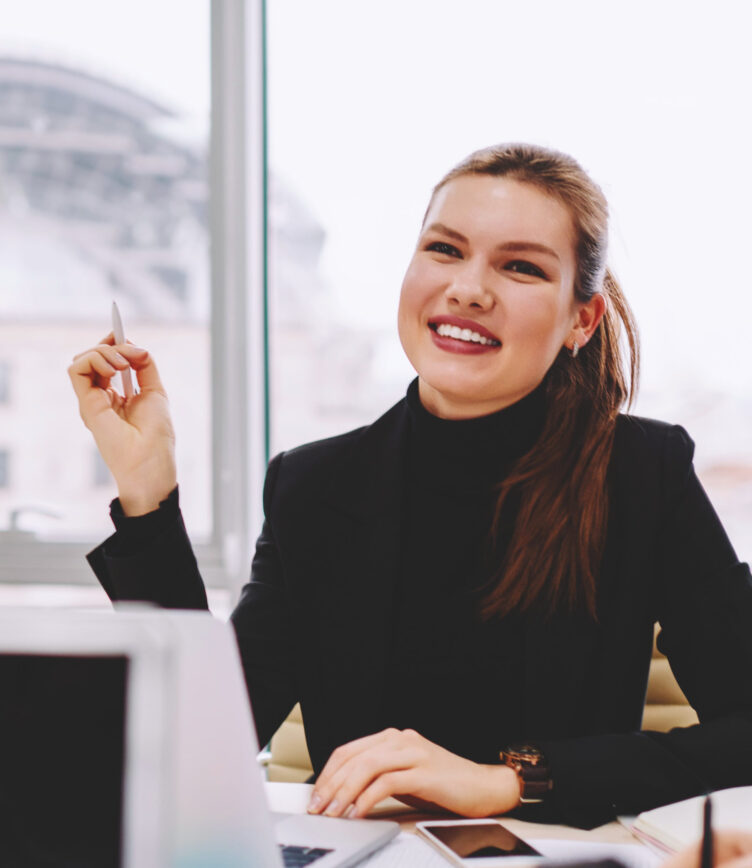Eine Frau im Business-Look sitzt in einem modernen Büro, lächelt und hält einen Stift, während sie in einer Besprechung ist.