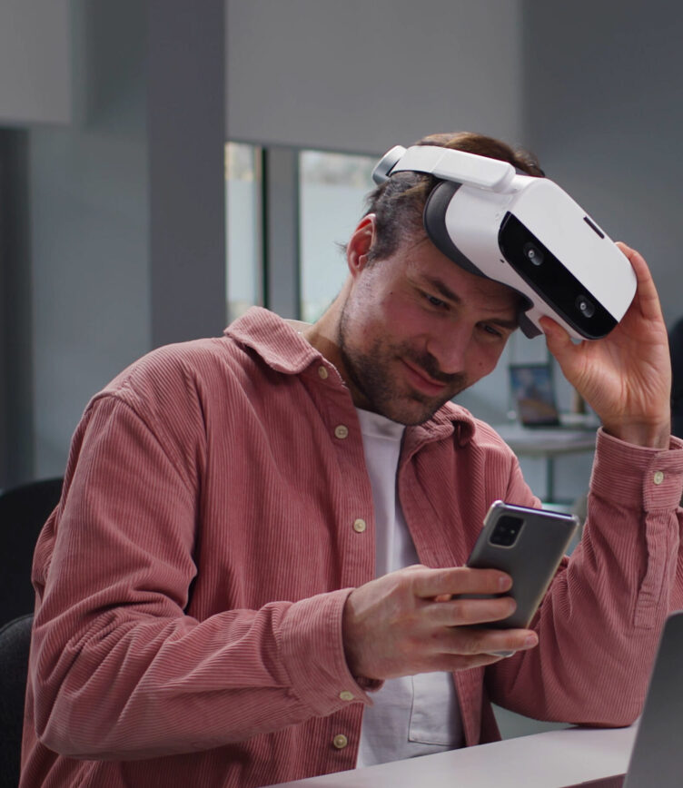 Eine Person trägt ein VR-Headset und hält ein Smartphone in der Hand. Die Person trägt ein rotes Hemd und sitzt an einem Tisch.