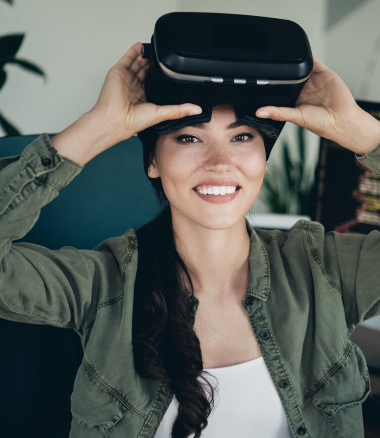 Eine Person trägt ein schwarzes Virtual-Reality-Headset und hält es mit beiden Händen. Sie trägt ein grünes Oberteil und sitzt in einem modernen Raum.
