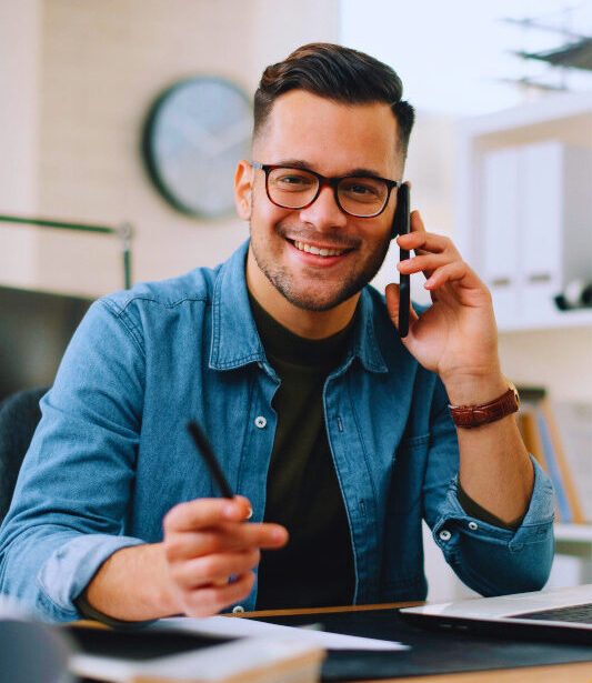 Ein junger Mann sitzt an einem Schreibtisch mit Laptop, Notizbuch und Kamera. Er trägt eine Brille und ein Jeanshemd, telefoniert mit dem Smartphone und lächelt in die Kamera. In der Hand hält er einen Stift, der Arbeitsplatz ist modern und hell eingerichtet.