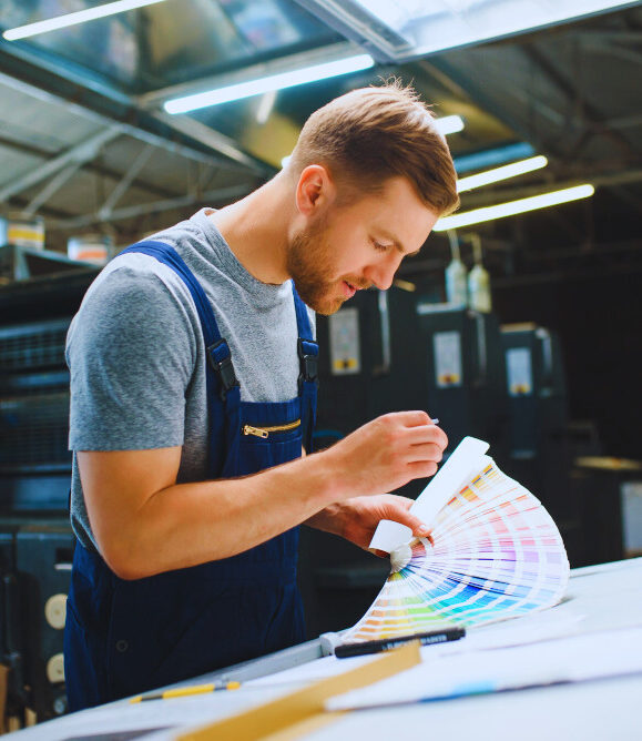 Ein junger Mann in Arbeitskleidung mit grauem T-Shirt und blauer Latzhose steht in einer Druckerei. Er betrachtet aufmerksam eine große Farbfächerpalette, um Farben zu prüfen. Im Hintergrund sind Druckmaschinen und technische Anlagen zu sehen.