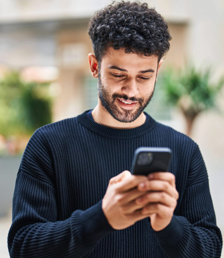 Junger Mann schaut lächelnd auf sein Smartphone und tippt eine Nachricht.