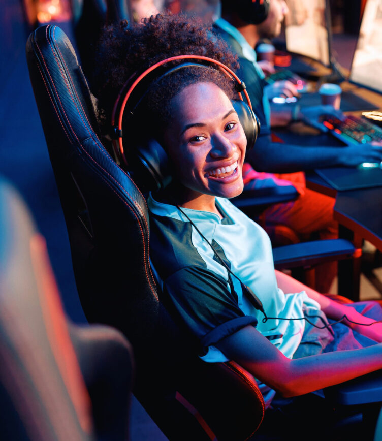 Junge Frau mit Headset sitzt in einem Gaming-Stuhl und lächelt in die Kamera, während im Hintergrund ein E-Sport-Turnier stattfindet.