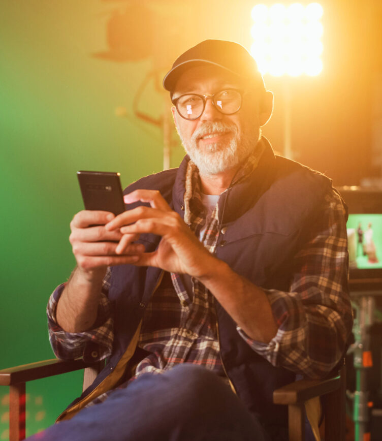 Älterer Mann mit Brille, Cap und Smartphone sitzt im Regiestuhl am Filmset mit starker Beleuchtung im Hintergrund.