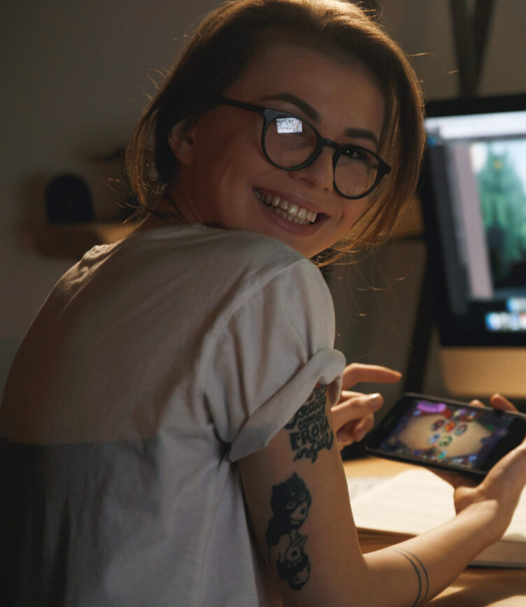 Junge Frau mit Brille und sichtbarem Tattoo lächelt, während sie ein Handyspiel spielt.