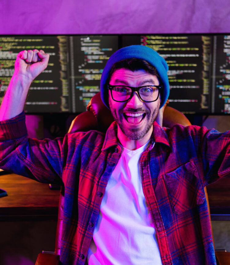 Begeisterter Programmierer mit Brille und Mütze sitzt vor mehreren Bildschirmen mit Code.