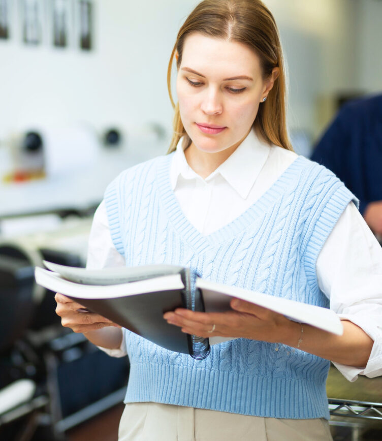 Eine Person hält zwei Bücher in den Händen, trägt einen blauen Strickpullover über einem weißen Hemd und steht in einem Raum mit Arbeitsplätzen im Hintergrund.