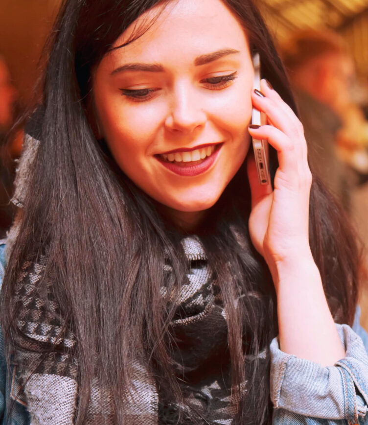 Eine Person mit langen, schwarzen Haaren hält ein Smartphone ans Ohr. Sie trägt eine graue Jeansjacke und einen schwarz-weißen Schal.