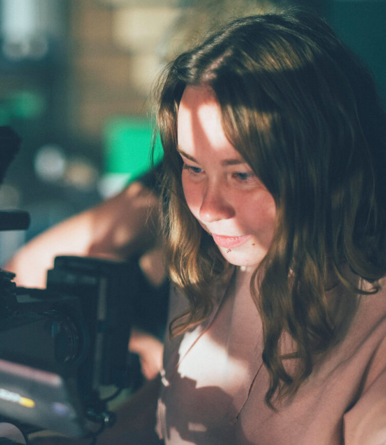 Eine junge Frau mit langen, braunen Haaren arbeitet konzentriert an einer Kamera oder technischem Equipment. Das Licht wirft Schattenmuster auf ihr Gesicht, was eine kreative und filmische Atmosphäre erzeugt.