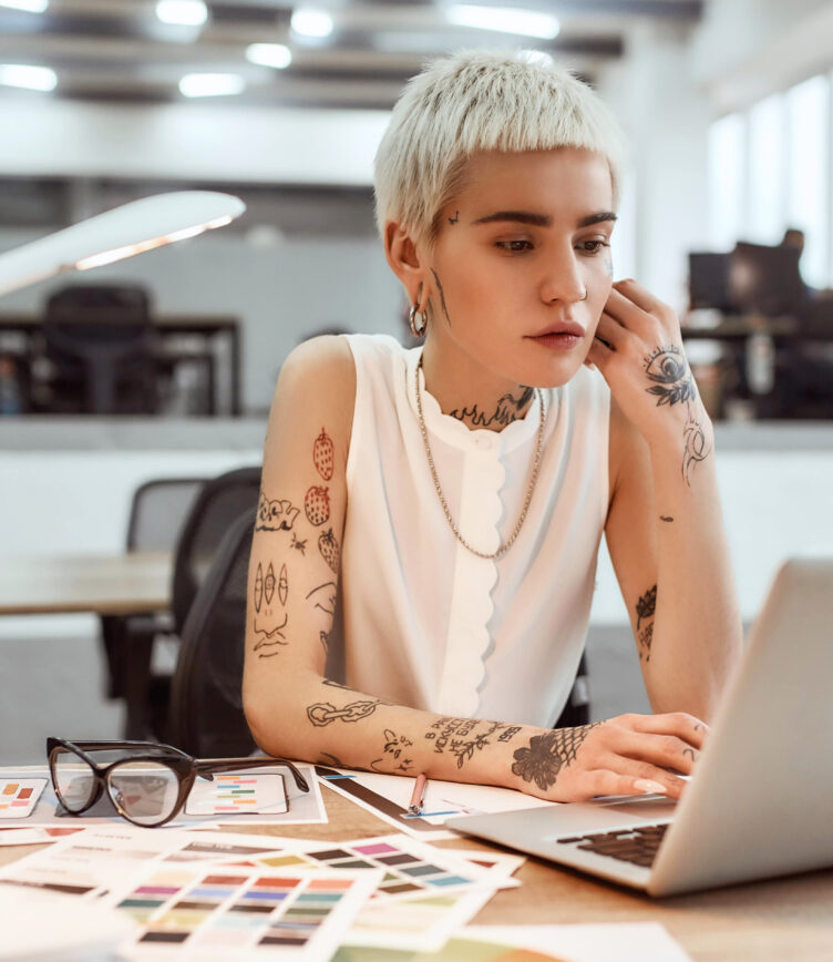 Eine Person mit kurzen, blonden Haaren und Tattoos sitzt an einem Tisch. Vor ihr liegen Farbproben und ein Laptop. Sie trägt ein ärmelloses Oberteil und hält eine Hand an ihr Gesicht.