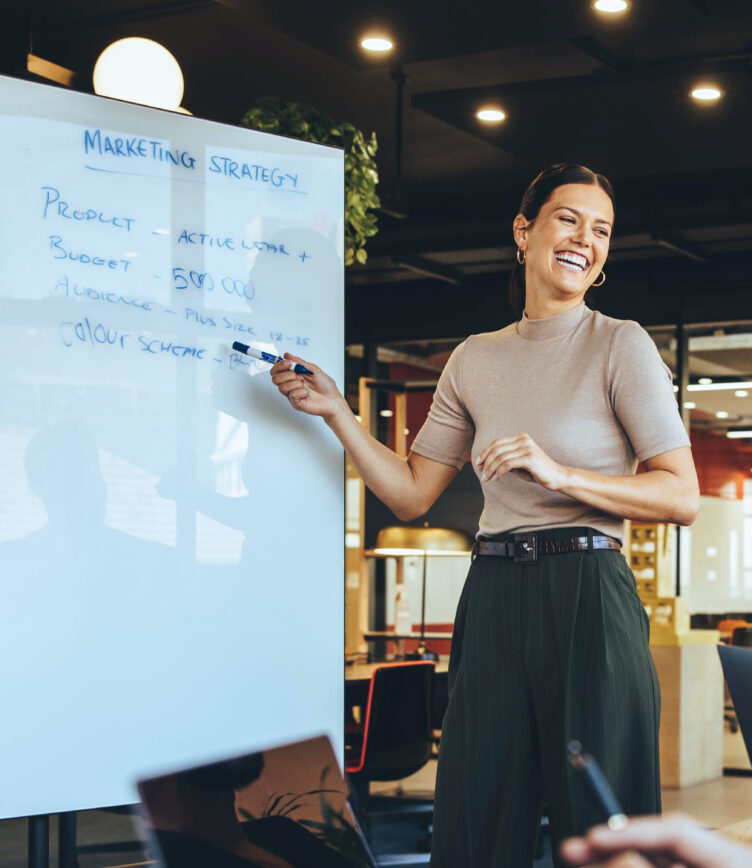 Frau mit Stift zeigt auf ein Whiteboard mit der Aufschrift 'Marketing Strategy'. Im Vordergrund sind Hände, die auf einem Tisch liegen.