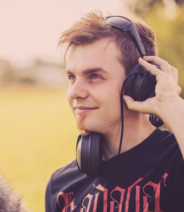 Junger Mann mit Kopfhörern, der mit der rechten Hand einen Kopfhörer an seinen Kopf hält. Er trägt ein schwarzes T-Shirt mit rotem Schriftzug.