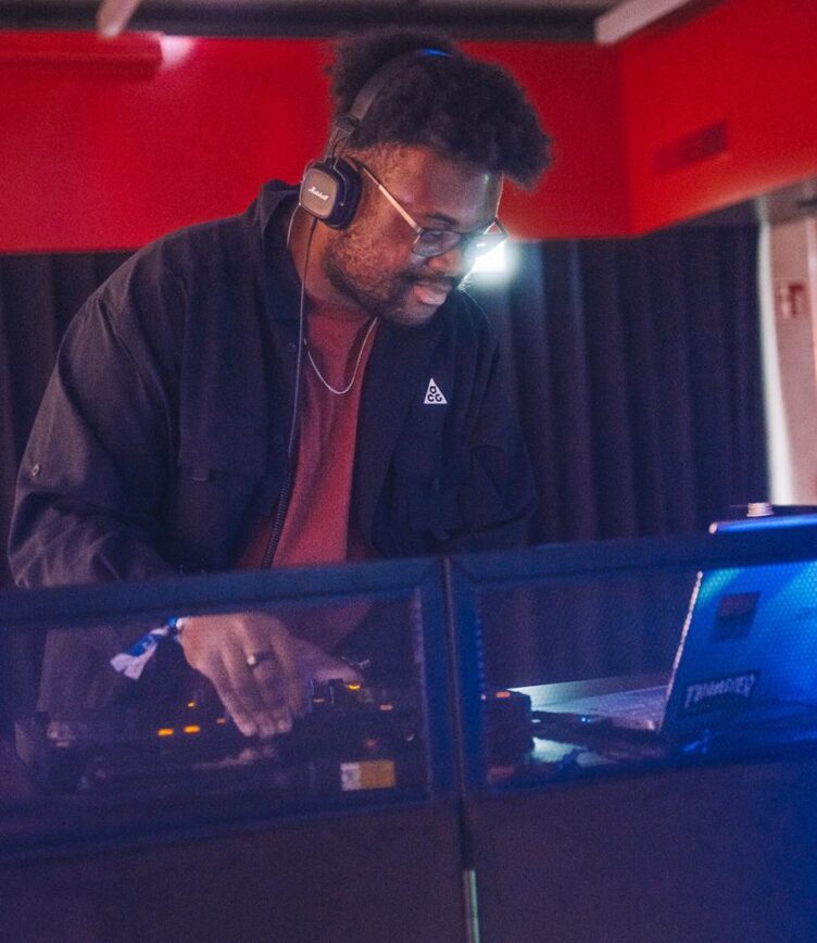 Ein DJ mit Kopfhörern steht an einem Mischpult in einem Club oder Veranstaltungsraum. Er trägt ein schwarzes Hemd über einem roten T-Shirt und konzentriert sich auf die Bedienung der Regler. Neben ihm steht ein Laptop mit Aufklebern, der für die Musiksteuerung genutzt wird.