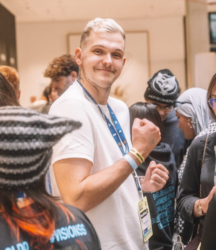 Ein junger Mann mit blondierten Haaren und weißem T-Shirt steht in einer Gruppe von Menschen bei einer Veranstaltung. Er lächelt in die Kamera, trägt ein Namensschild und ein blaues Lanyard um den Hals. Um ihn herum unterhalten sich andere Teilnehmende, einige halten Getränkeflaschen in der Hand.