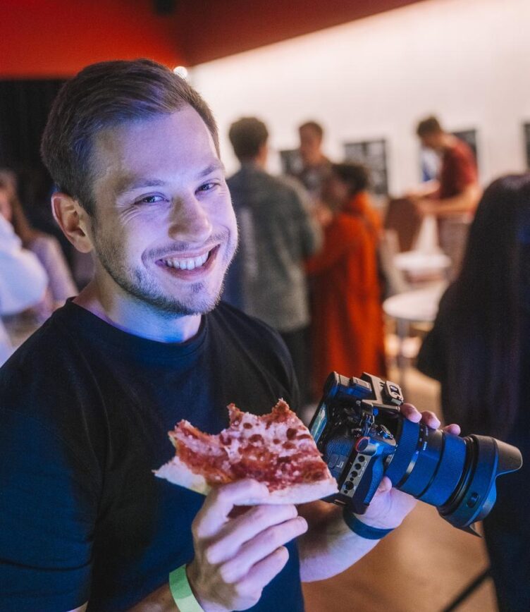 Ein junger Mann lächelt in die Kamera, hält ein Stück Pizza in der einen Hand und eine große Kamera in der anderen. Im Hintergrund sind weitere Personen bei einer Veranstaltung zu sehen.