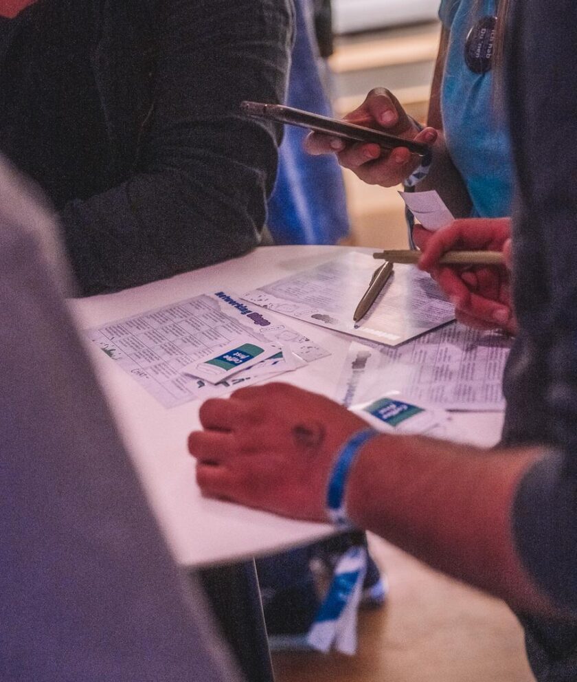 Mehrere Personen stehen um einen Tisch und füllen Formulare aus. Auf dem Tisch liegen Stifte, Zettel und Flyer mit dem Logo „Start into Media“. Eine Person hält ein Smartphone in der Hand, eine andere trägt ein blaues Festivalband am Handgelenk.