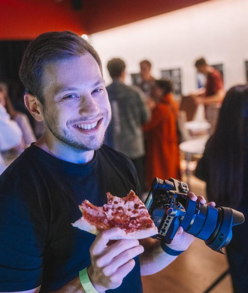 Ein junger Mann lächelt in die Kamera, hält ein Stück Pizza in der einen Hand und eine große Kamera in der anderen. Im Hintergrund sind weitere Personen bei einer Veranstaltung zu sehen.