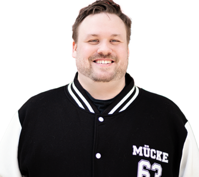 Fröhlicher Mann mit kurzem Haar und Bart, der eine schwarze College-Jacke mit weißen Ärmeln trägt. Auf der Jacke steht „Mücke 63“.