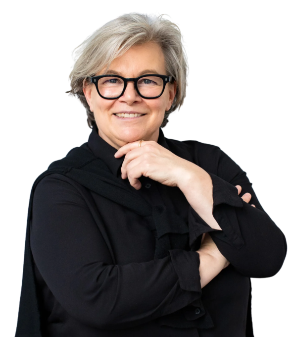 Ältere Frau mit kurzen, grauen Haaren und schwarzer Brille, die lächelnd in die Kamera schaut. Sie trägt eine schwarze Bluse und hat eine Hand unter ihr Kinn gelegt, während die andere verschränkt ist.