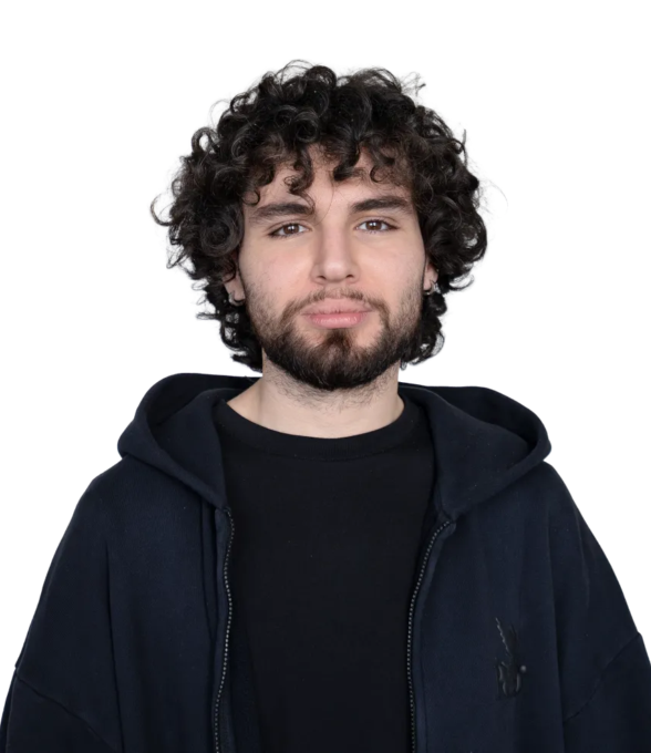 Junger Mann mit lockigem, dunklem Haar und Bart, der in die Kamera schaut. Er trägt einen schwarzen Hoodie über einem schwarzen T-Shirt.