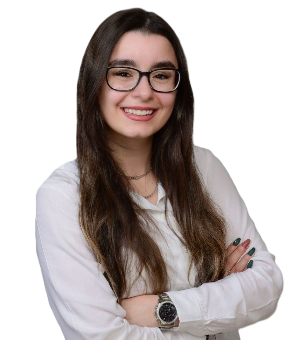 Junge Frau mit langen, dunkelbraunen Haaren und schwarzen Brillengestell, die lächelnd in die Kamera schaut. Sie trägt eine weiße Bluse und eine Armbanduhr und hat die Arme vor der Brust verschränkt.