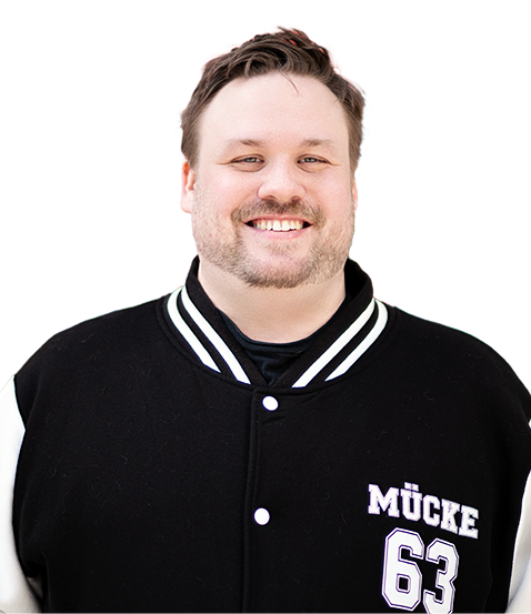 Fröhlicher Mann mit kurzem Haar und Bart, der eine schwarze College-Jacke mit weißen Ärmeln trägt. Auf der Jacke steht „Mücke 63“.