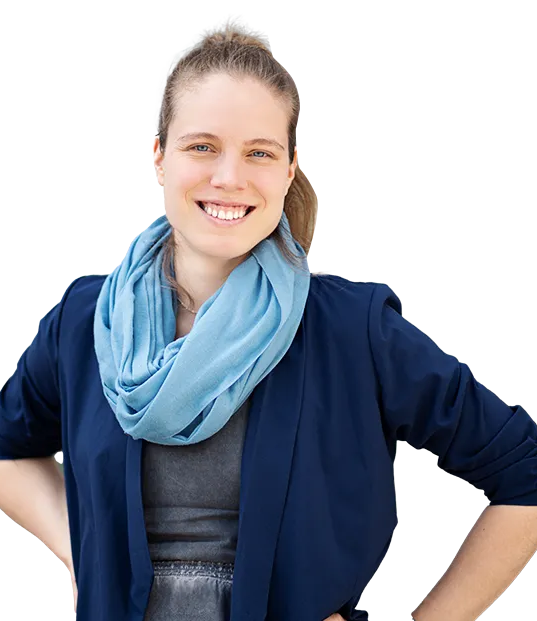 Junge Frau mit blondem, zurückgebundenem Haar, die lächelnd in die Kamera schaut. Sie trägt ein dunkelblaues Jackett, einen hellblauen Schal und ein graues Oberteil. Ihre Hände sind in die Hüften gestützt.