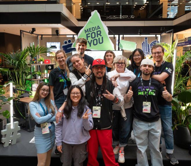 Eine größere Gruppe junger und älterer Menschen posiert gut gelaunt für ein Gruppenfoto auf einem Event. Im Hintergrund stehen Pflanzen, bunte Deko und ein großer Schriftzug „MEDIA FOR YOU“. Viele der Personen tragen Event-Badges und schwarze T-Shirts mit Medien-Motiven und machen fröhliche Posen oder Handzeichen.
