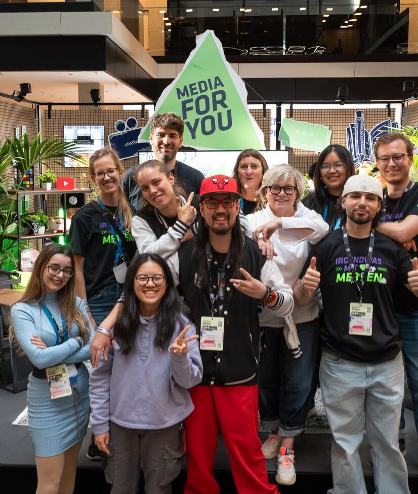 Eine größere Gruppe junger und älterer Menschen posiert gut gelaunt für ein Gruppenfoto auf einem Event. Im Hintergrund stehen Pflanzen, bunte Deko und ein großer Schriftzug „MEDIA FOR YOU“. Viele der Personen tragen Event-Badges und schwarze T-Shirts mit Medien-Motiven und machen fröhliche Posen oder Handzeichen.
