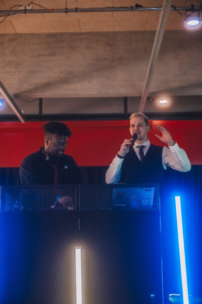 Zwei Männer stehen auf einer Bühne hinter einem Mischpult. Der rechte mit blondierten Haaren und in einem Anzug spricht in ein Mikrofon, der linke bedient das DJ-Pult. Blaue Leuchtstäbe am Pult ergänzen die Lichtstimmung.