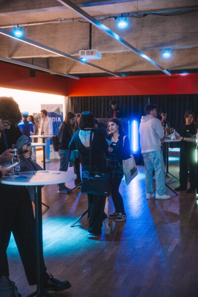 In einer kleinen Halle mit warmer Lichtstimmung sind mehrere Personen sind um Stehtische herum versammelt und führen Gespräche. Einige von ihnen halten Flyer und Karten.