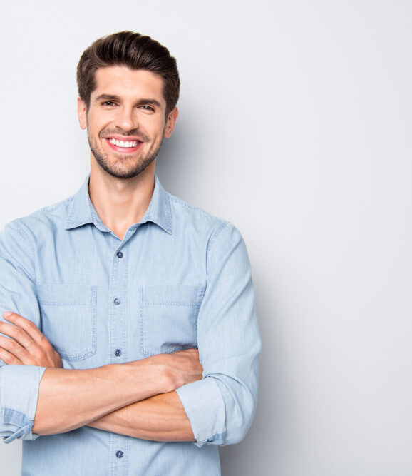 Junger Mann mit blonden Haaren in einem lockeren, hellblauen Hemd steht selbstbewusst und sympathisch lächelnd vor einer neutralen Wand.