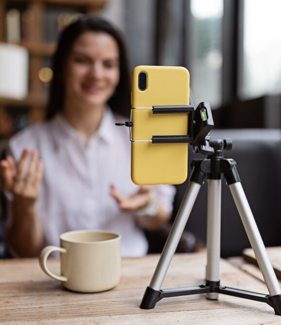 Im Vordergrund ist ein gelbes Smartphone in einem kleinen Stativ zu sehen, das auf dem Tisch aufgestellt wurde. Im Hintergrund etwas verschwommen eine weibliche Person mit braunen Haaren und einem weißen Hemd. Neben dem Stativ auf dem Tisch steht eine beige Tasse.
