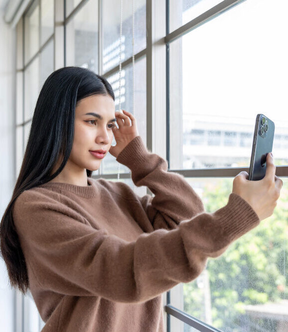 Eine junge Frau mit schwarzen Haaren und einem braunen Pullover steht vor einer Fensterwand mit viel natürlichem Licht und betrachtet ihr Bild im Smartphone.