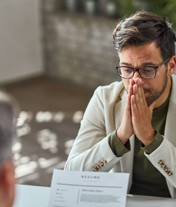 Junger Mann mit Brille sitzt in einem hellen Anzug beim Bewerbungsgespräch. Er sieht besorgt aus, während sein Gesprächspartner ein Papier mit der Aufschrift "Resume" liest.