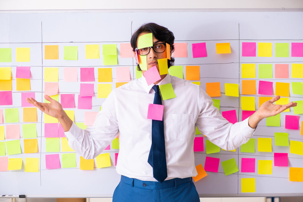 Mann steht vor einem Whiteboard voller bunter Post-its, viele Zettel kleben auch an seinem Gesicht und Anzug – Symbol für Chaos oder Überforderung im agilen Arbeiten.