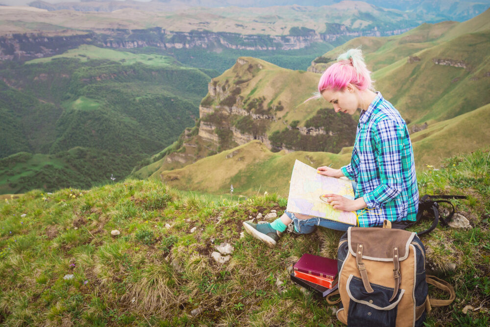Junge Frau mit pink gefärbten Haaren sitzt mit Karte und Rucksack auf einer Bergwiese und studiert den Weg – im Hintergrund eine weitläufige Landschaft.