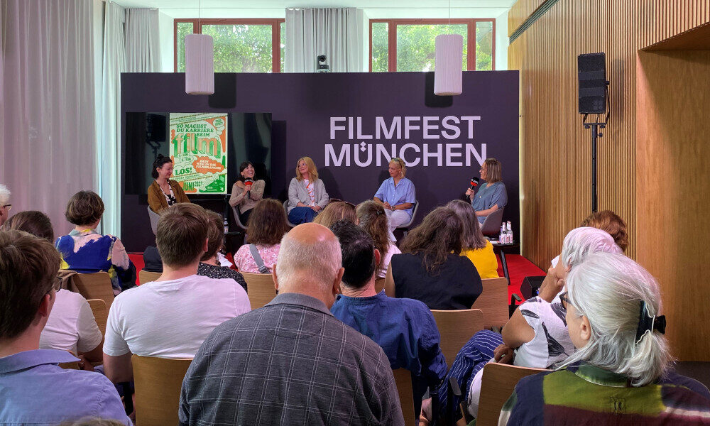 Podiumsdiskussion beim Filmfest München: Fünf Panelist:innen sitzen auf einer Bühne vor einem Publikum, im Hintergrund ist ein großes Logo „FILMFEST MÜNCHEN“ zu sehen.
