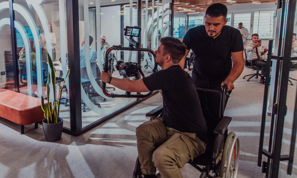„Ein Kameramann im Rollstuhl wird von einem Kollegen durch ein modernes Büro geschoben, während er eine Filmszene aufnimmt.“