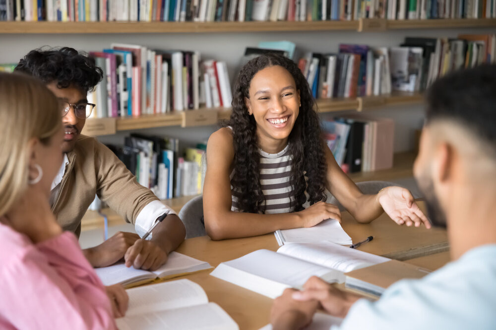 „Gruppe junger Menschen arbeitet zusammen in einer Bibliothek, eine lachende Frau spricht angeregt mit ihren Mitstudierenden.“